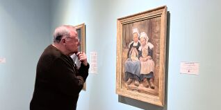A man leans in as he views a painting of a mother and a daughter. The painting is in a gold frame and hangs on a light blue wall.